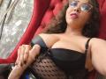 BarbaraLeythonn with curly brown hair, glasses, and pink lipstick, lying in a red chair, wearing a black bra with mesh and bow, holding a large, uncircumcised, erect penis.