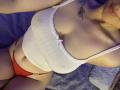 Olieri's white, ribbed, sleeveless top with a small bow, covering her tits, and red panties, sitting on a bed. Tattoo of leaves on her right shoulder.