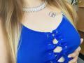 A blusa cropped azul com textura em tiras da KarKutie, com amarração na frente, que mostra seus seios grandes, tem um colar de renda prateado e uma tatuagem no peito. Seu cabelo loiro está solto..