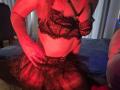 Milagres with red-lit skin, wearing black lace bra and sheer skirt, showing her tits and hand on her ass. Kneeling on blue bed, background blurred.