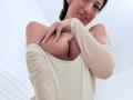 Mireille Veil, avec de grandes seins, souriante, portant un haut blanc moulant, dégagé sur une épaule, et cachant ses mamelons avec sa main, une bague visible à son doigt, cheveux foncés et peau pâle..