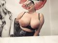 GeorgiaLerox with huge tits, black bra, and glasses, leaning forward in a leather chair, black hair in a bun, tattoo on right arm, red lipstick, white and black background.