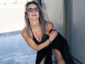 Milagres with wet purple hair, black dress, and sunglasses, holding pink vibrator, smiling, standing by a wall, wearing choker, and black bracelet.