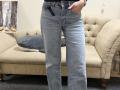 Dalaila's ass is covered by light blue, slightly flared jeans with a button fly and a black belt, standing in front of a beige tufted sofa.
