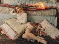 KylaHemeke, with wavy blonde hair, wearing a white dress and pearl necklace, sits on a dark floor, legs crossed, surrounded by frilled pillows and a lit candelabrum.