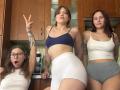 Three HotCaramelsTrio women in a kitchen: one with glasses, open mouth, white tank, white shorts; one with blue bra, white shorts, tattooed arm, peace sign; one in white tank.