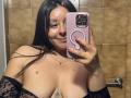 EmaJames with long black hair, holding a pink phone, smiling, topless with large boobs, wearing a black lace sleeve, and a necklace, in a bathroom.