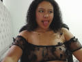 AriaVega with curly black hair, tongue out, wearing a sheer black off-shoulder top, showing small boobs, and a gold necklace, in a bedroom.