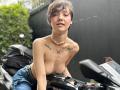 LissyLuxe with short, dark hair, topless, small tits, butterfly tattoos, necklace, jeans, leaning on a black scooter, black bag in background, green bush on left.