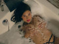 LindaJimenex with dark hair, red lipstick, and wet skin, lying in a bathtub full of bubbles, covering her tits with hands, wearing black panties, looking up.