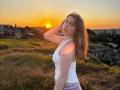 LeScharwath with wavy brown hair, white tank top, and purple pants, stands on a grassy hill, hand in hair, sunset glowing behind her, sun lens flare on her shoulder.