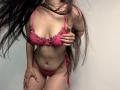 Tisia with long dark hair, wearing pink lace bra and panties with floral accents, has hand on chest, showing her tits and pussy, standing with hand on thigh.