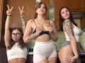 Three HotCaramelsTrio standing in a kitchen, wearing white and blue sports bras, and high-waisted white shorts. One has tattoos, another is making a double peace sign.