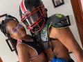 LilithAndAsmodeo with braided hair, glasses, and black sports bra, standing next to LilithAndAsmodeo in football gear, red helmet, black shoulder pads, showing black bra and tattoo.