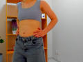 Steisii with a flat, toned stomach, wearing a blue sports bra and high-waisted denim jeans, standing with hands on hips, showing a tattoo on the right side.
