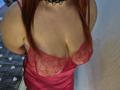 Dinastiia with red lace bralette, showing big boobs, in a pink satin dress, black lace choker, red hair, and fair skin, standing in a doorway.