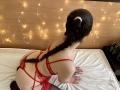 Tisia has a long braid, red lace lingerie, and garter, kneeling on a bed, showing her butt and back, with fairy lights in the background.