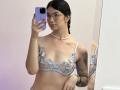 EvaaFarix with glasses, black hair, wearing a sheer blue floral bralette, necklace, and bracelet, holding a purple phone, mirror selfie, visible tattoo on right arm.