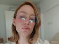 MissFrenchy with pinkish lips, light skin, and shoulder-length blonde hair, wearing round gold-rimmed glasses, showing a reflection of a blue heart on the glass.