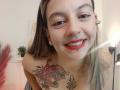 ArianaBlonde with red lipstick, pierced lower lip, and tattoos on her chest, including a red rose and black leaf, smiling at the camera, wearing a headband.