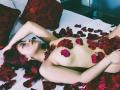 ShofiaWett with red hair, lying topless on a bed, covered in red rose petals, arms above head, looking up, white bed, red floral pillows, silver bracelet, necklace.