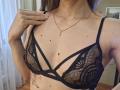 MilaYanis with her tits in a black, lace, strappy bra, showing her small moles, tattoo on her right side, and a gold necklace, hand on her collarbone.