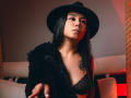 MissCharlotteJolie with long black hair, wearing a black hat, lace bra, and fuzzy black jacket, reaching forward with a ring, red light in background, tattoo on right chest.