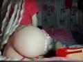 Madiy69 with platinum blonde hair, red top, and pink lace panties, showing her big ass and tattoo, kneeling on a bed with red heels and a white pillow.