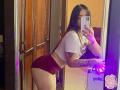 SophiaMarx with long brown hair, white crop top, and red velvet butt shorts, leaning on a mirror, showing tattoos on her right arm, holding a phone, purple light.