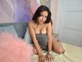 RubyCrow with dark hair, sitting on a bed, wearing a gray strapless top, showing her tits, pink fluffy pillow, white watch, and a subtle smile, in a cozy room.