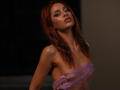 JesicaRare with red hair, sultry expression, and purple sheer wrap covering her tits, standing in dim light. Her skin is shiny, and she's looking slightly up.