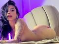 Kazumii is lying naked on a couch, her tattooed arm in front, butt up, black hair covering one eye, and wearing gold hoop earrings, with purple lighting.