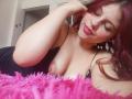 SophieHill with red hair, nose ring, and pink lipstick, lying on a pink fluffy blanket, showing her tits, eyes closed, black dress, hand near face, smiling.