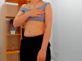 Steisii with a light gray off-shoulder top, black skirt, hand covering small tits, silver necklace, visible navel, light skin, standing in a room with wooden shelves.