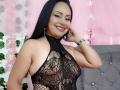 Zaria69 with long black hair, smiling, wearing a black, lace, halter top that reveals her tits and has a netted pattern. She's touching her face with her right hand.