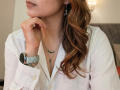 AnaEoxo with wavy brown hair, white button-down shirt, green necklace, silver cross earrings, and a light blue watch, touching her chin. Boobs slightly visible under shirt.