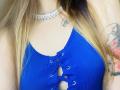A blusa cropped azul com textura em tiras da KarKutie, com amarração na frente, que mostra seus seios, possui um colar com diamantes e tatuagens no ombro. Ela tem cabelo loiro e pele clara..