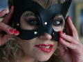 CandyLadyZ with a black mask, blue eyes, red lipstick, and red nails, holding the mask to her face. Her tits are slightly visible, and her pink lips are parted.
