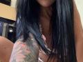 AndraD with long black hair, showing a tattooed arm and pink bra, her tits slightly visible, sitting with legs apart, focusing on her upper body.