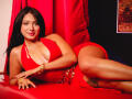 LilyConorr with dark hair, red halter dress, and gold bracelets, lying on red leather, showing her tits and ass, smiling, legs crossed, and tattoo on her right thigh.