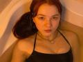 KatiRokisky with red hair, looking up, in a bathtub, wearing a black halter top, showing small tits, water covering her chest, and a chain necklace.