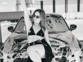 Milagres with tits in a black bikini top, dark skirt, and sunglasses, sitting on a soapy car, raising one arm, in a black-and-white photo.