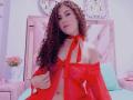 IrisDalton with curly brown hair, wearing a sheer red off-shoulder top with ribbon, exposing her tits and a small red bow on her chest, sitting on a bed.