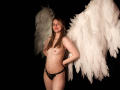 AmberWoody with white angel wings, standing naked, wearing black lace panties, hands behind back, pale skin, small tits, brown hair, red lipstick, black background, white feathers.