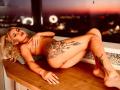 NayaBliss is lying naked on a wooden windowsill, covering her tits with one hand, showing off large tattoos on her thigh and leg, with a city skyline in the background.