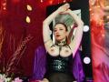 QueenScarletSecret with arms raised, wearing black leather harness exposing her tits, standing in front of mirror, red curtain background, purple chair, and string lights.
