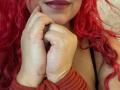 Estefy69's red curly hair, hands touching her chin, wearing a red ribbed sleeve and dark bra, with glossy purple lipstick. Her tits are visible but not the focus.