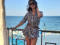 MiraMixi with black hair, sunglasses, and a leopard-print dress, brown belt, standing by a balcony, ocean in background, holding her hair, looking sexy, and showing her ass.