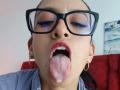 ZarayFeet with glasses, tongue out, and drooling, has pink lips, red checkered shirt, gold hoop earrings, and visible moles on her face. Her tongue is covered in white fluid.