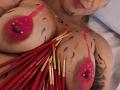 DahianaDmour's tits are painted with pink and red splotches, nipple piercings, and multiple red paintbrushes are stuck to her chest. She has a rose tattoo on her side.